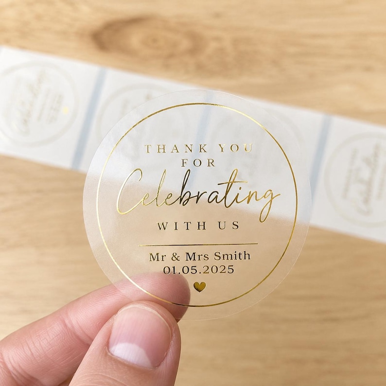 Hand holding a clear round wedding thank you sticker with metallic gold foil text reading “Thank You for Celebrating With Us” on a wood background, featuring personalised names and wedding date with matching labels blurred behind.
