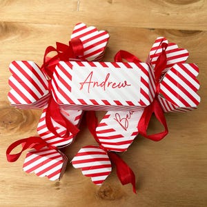 Puede incluir: Un conjunto de cajas de regalo con forma de caramelo a rayas rojas y blancas con lazos rojos. Las cajas están decoradas con los nombres "Andrew" y "Bea" en escritura roja. Dispuestas sobre una superficie de madera.
