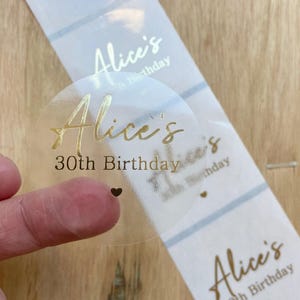 May include: Clear, round sticker with gold foil text that reads "Alice's 30th Birthday". The sticker is on a roll of similar stickers with a light blue and white striped background. A small black heart is visible.