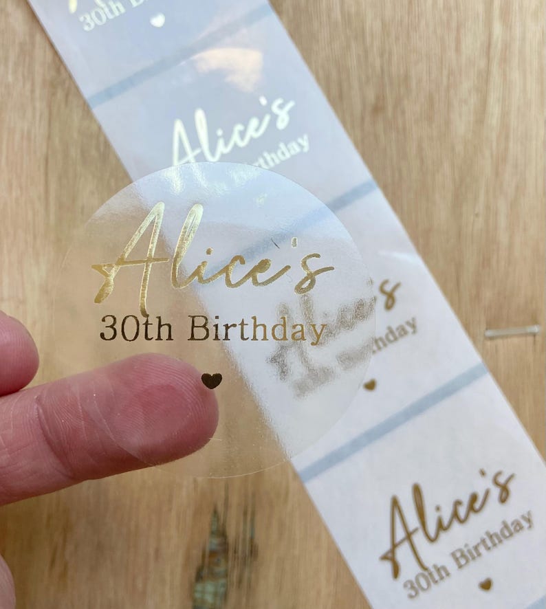 May include: Clear, round sticker with gold foil text that reads "Alice's 30th Birthday". The sticker is on a roll of similar stickers with a light blue and white striped background. A small black heart is visible.