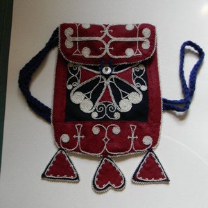 Op de afbeelding: Een rode en zwarte kralen tas met een blauwe band en een witte knoop. De tas heeft een vlinder ontwerp en is versierd met witte kralen.