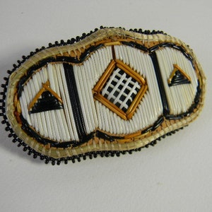 Peut inclure: Barrette ovale faite à la main, ornée de perles. Elle présente un motif central en losange et deux triangles, tous en noir, blanc et or. Les bords sont soulignés de perles noires et d'une matière tissée semblable à de la paille.