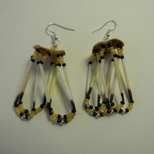 May include: A pair of handcrafted earrings with a distinctive design. They feature tan leather tops with silver hooks. Below, multiple off-white quills with dark brown tips are accented with black and yellow beads, creating a unique, artistic appearance.
