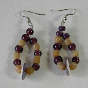 May include: A pair of handmade earrings with a teardrop design. The earrings feature a combination of dark purple and light yellow beads, with a small, flat, white accent piece at the bottom. The ear wires are silver-colored.