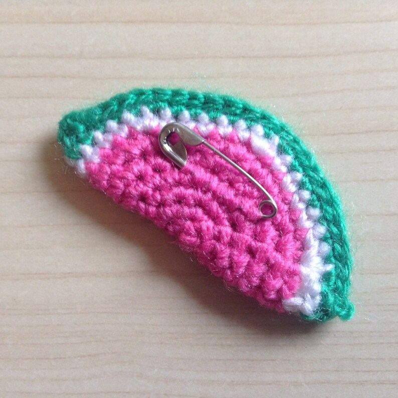 May include: A crocheted watermelon slice with a green and white border and a pink center. A safety pin is attached to the back.