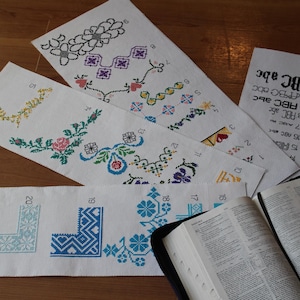 May include: Several cross-stitch samplers with floral and geometric designs in various colors, including blue, pink, and yellow. A Bible is open next to the samplers, and a sheet with the alphabet is also present.