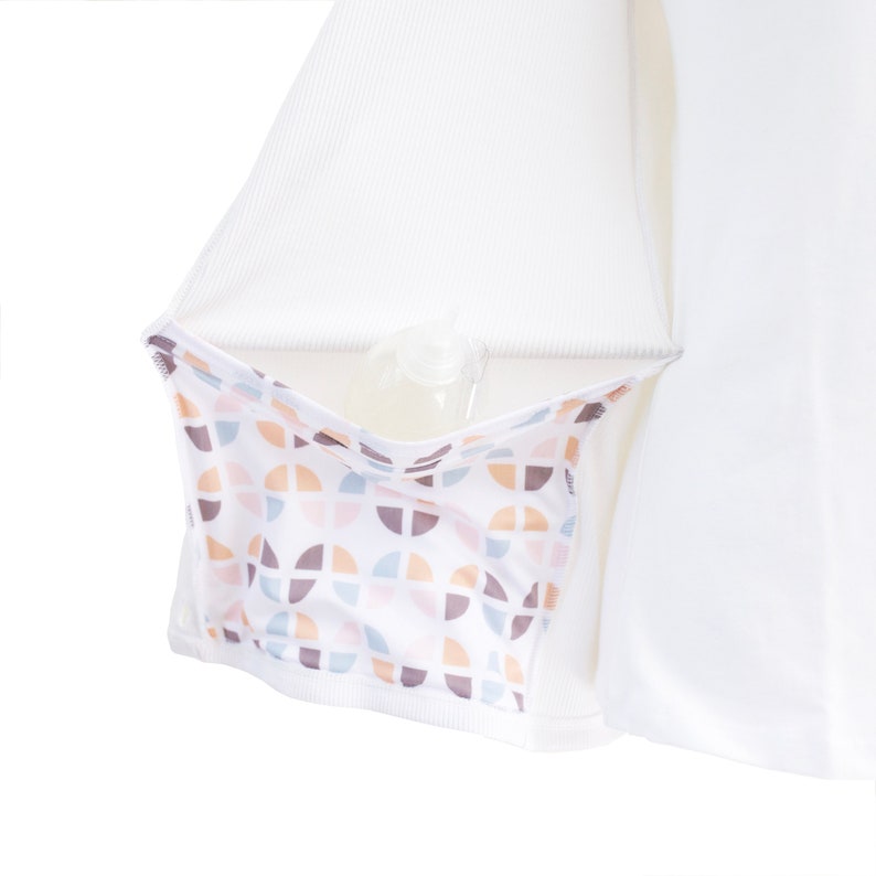 Puede incluir: Camiseta blanca de manga larga con un bolsillo oculto que tiene un patr&oacute;n geom&eacute;trico en tonos de rosa, marr&oacute;n y azul. El bolsillo est&aacute; dise&ntilde;ado para guardar una botella peque&ntilde;a u otros art&iacute;culos.