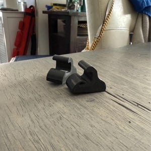 May include: Two black plastic clips, shaped like a hook, are on a wooden surface. The clips are designed to hold items in place.