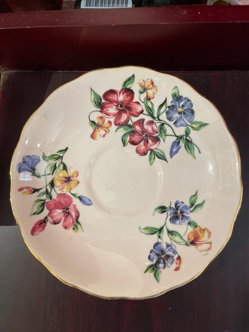 May include: A white saucer with a gold rim and a floral design. The flowers are pink, red, blue, and yellow.