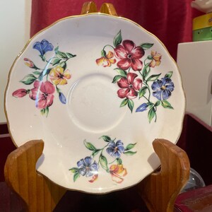 May include: A white porcelain saucer with a gold rim and a floral design featuring pink, yellow, blue, and green flowers. The saucer is on a wooden stand.