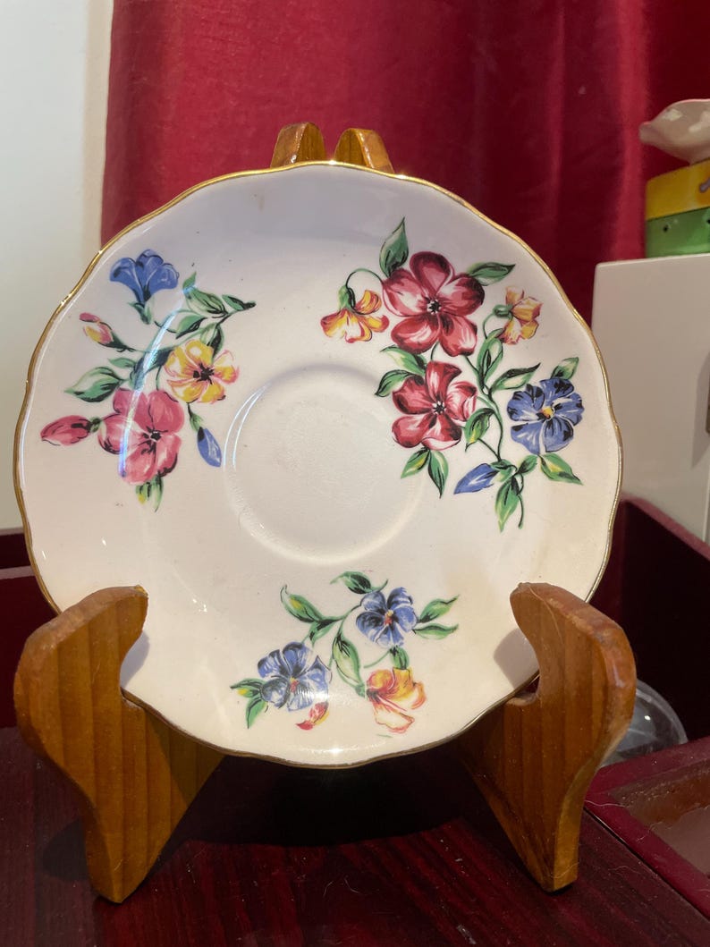 May include: A white porcelain saucer with a gold rim and a floral design featuring pink, yellow, blue, and green flowers. The saucer is on a wooden stand.