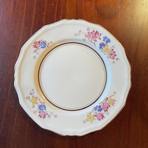 May include: A white ceramic plate with a scalloped edge and a gold rim. The plate is decorated with a floral pattern in shades of pink, yellow, and blue.