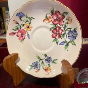 May include: A white saucer with a gold rim and a floral pattern in shades of pink, blue, yellow, and green. The saucer is on a wooden stand.