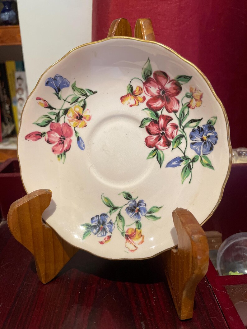 May include: A white saucer with a gold rim and a floral pattern in shades of pink, blue, yellow, and green. The saucer is on a wooden stand.
