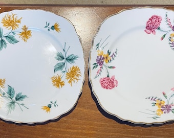 May include: Two white ceramic plates with gold trim and floral designs. One plate features yellow flowers and green leaves, while the other has pink, purple, and yellow flowers.