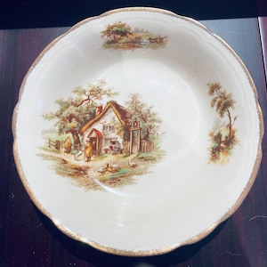 Puede incluir: Un tazón de cerámica blanca con un borde dorado y una escena de campo pintada en el frente. La escena presenta una pequeña cabaña con techo de paja, árboles y personas caminando por un sendero.