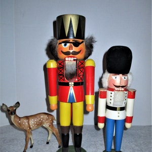 May include: Two wooden nutcrackers, one large and one small, stand side-by-side. The larger nutcracker wears a red and yellow uniform with a black hat. The smaller nutcracker wears a white and blue uniform. A small deer figurine stands to the left.