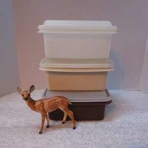 May include: A stack of three rectangular plastic storage containers with lids, featuring a clear top container, a beige middle container, and a dark brown bottom container, accompanied by a small brown and white spotted deer figurine.
