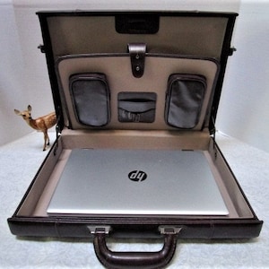 May include: Open brown leather briefcase with a silver HP laptop inside. The interior features a tan lining and several pockets. A small deer figurine stands to the left of the briefcase.