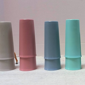 May include: A collection of six pastel-colored plastic cups in shades of gray, pink, blue, and green. The cups are stacked, with the top portion inverted on the bottom. A small deer figurine is in the background.
