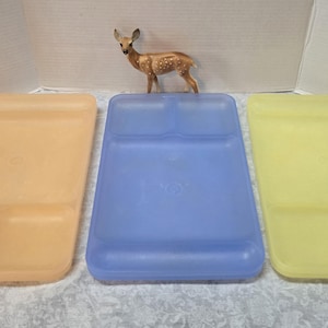 May include: Three vintage plastic divided plates in shades of yellow, blue, and orange. Each plate has a rectangular shape with two or three compartments. A small deer figurine stands on top of the blue plate.