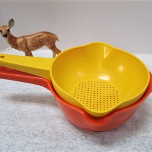 May include: Two plastic colanders, one yellow and one orange, nested together. The yellow colander has a strainer bottom and a handle. The orange colander has a smooth bottom.