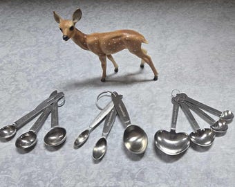 May include: A set of eight stainless steel measuring spoons. The spoons are arranged in two groups of four. The spoons are all different sizes and have a variety of shapes, including round, heart, and oval.