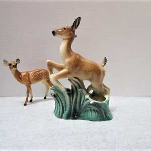 May include: Two ceramic deer figurines. One deer is leaping from a teal-colored base with green grass accents. The other deer is standing. Both deer are tan with white spots and black accents.