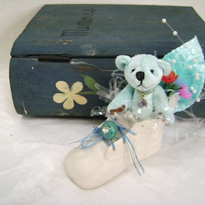 May include: A teal teddy bear with a jeweled necklace and a decorative wing sits in a white ceramic baby shoe. The shoe is adorned with a blue ribbon and a green jewel. A blue wooden box with the word "Memories" is in the background.