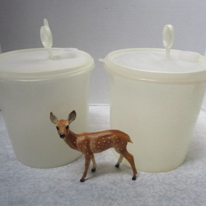 May include: Two off-white plastic containers with lids and a small, brown deer figurine with white spots. The containers are cylindrical with a handle on the lid. The deer is standing in front of the containers.