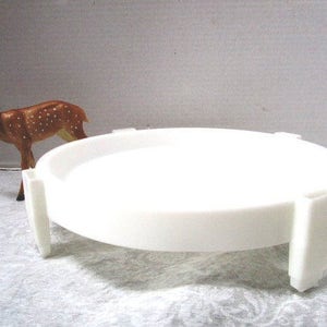 May include: White plastic cake stand with a round top and four legs. The stand is approximately 12 inches in diameter.