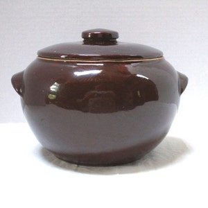 May include: A brown ceramic crock pot with a lid. The crock pot has two handles on the sides and a smooth, glossy finish.