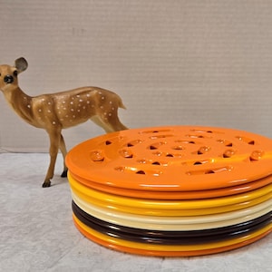 May include: A stack of colorful, round ceramic trivets in shades of orange, yellow, white, and brown. The top trivet is orange with a series of holes. A small deer figurine stands nearby.