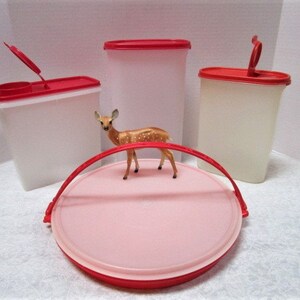 May include: A collection of vintage Tupperware containers in various sizes, featuring white bodies and bright red lids. One container has an open lid. A small deer figurine rests on a pink and red serving tray.