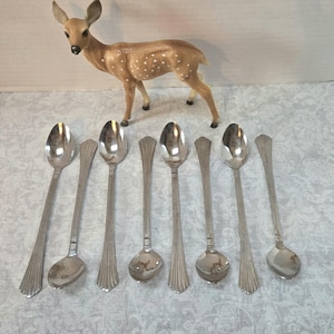 May include: A collection of seven silver spoons with a decorative handle design. A small, brown deer figurine with white spots is in the background. The spoons are arranged in a row on a patterned surface.