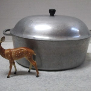 May include: A silver aluminum oval Dutch oven with a lid and two handles. The oven is sitting on a white surface with a small brown and white deer figurine in front of it.
