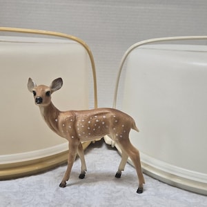 May include: Two off-white cake carriers with handles and a small, brown deer figurine with white spots. The cake carriers have a rounded shape and a light yellow trim. The deer is standing between the carriers.