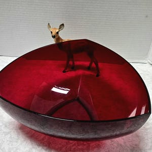May include: A deep red glass bowl with a triangular shape and rounded corners. A small, brown and white deer figurine stands inside the bowl, positioned toward the back edge. The glass is translucent, showing a smooth, polished surface throughout.