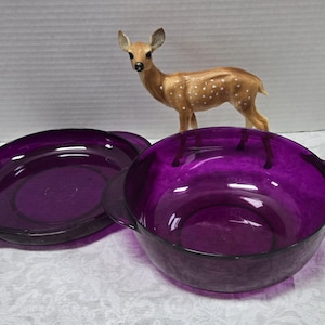 May include: Two deep, purple glass bowls with handles and a matching plate. A small, tan deer figurine with white spots stands behind the bowls. The bowls and plate are made of a shiny, transparent glass.