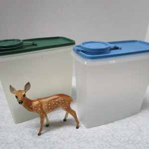 May include: Two clear plastic containers with lids. The container on the left has a green lid and the container on the right has a blue lid. A small, white deer figurine is in front of the containers.