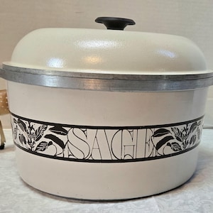 May include: Cream-colored vintage pot with lid and black handle. The pot has a silver rim and a black and white floral design with the word "SAGE" in the center. A small deer figurine is to the left of the pot.
