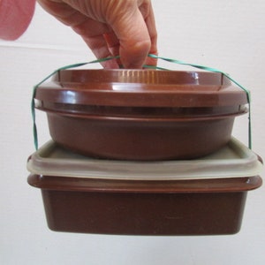 May include: Two brown plastic containers with lids, one stacked on top of the other, are held together by a green rubber band.