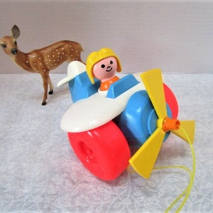 May include: A vintage toy airplane with a yellow propeller, blue and white body, and red wheels. A small plastic figure sits in the cockpit. A brown deer figurine stands nearby.