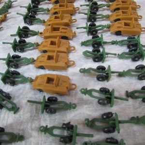 May include: A collection of small, tan toy tanks and green artillery pieces are arranged on a white surface. The toy tanks have a rectangular shape with small wheels, while the artillery pieces have a gun carriage design.