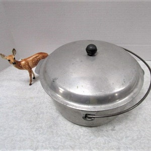 May include: A vintage aluminum cooking pot with a lid and a black handle. The pot has a rounded shape and a black knob on top of the lid. A small, brown deer figurine with white spots is next to the pot.