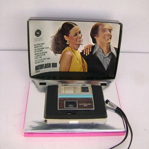 May include: A vintage black and silver Autoflash 100 camera with a black wrist strap. The camera is on a silver surface, with a box featuring a smiling couple. The box reads "AUTOFLASH 100 CAMERA".