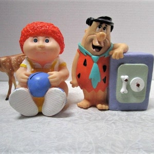 May include: A set of three vintage plastic figurines. A red-haired Cabbage Patch Kid doll holding a blue ball, a cartoon character resembling Fred Flintstone holding a safe, and a brown deer figurine.
