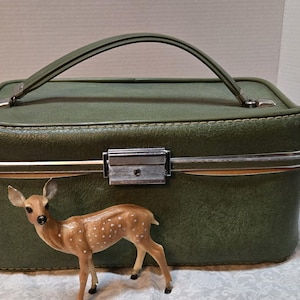 May include: A vintage green travel case with a silver clasp and handle. A small, brown deer figurine with white spots stands in front of the case. The case has a rectangular shape and a stitched design.