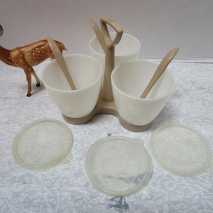 May include: A white ceramic condiment set with three individual bowls and a central stand. The set includes three spoons and is designed to hold condiments like salt, pepper, and sugar.