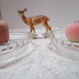 May include: Two clear glass pie plates with a decorative rim, one with a red apple and the other with a gold apple. A spotted fawn figurine is in the center of the image.